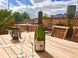 En flaske mousserende vin og to tomme champagneglas står på et udendørs træbord, mens folk spiser i baggrunden under en delvis overskyet himmel.
