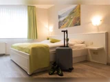 Et pænt hotelværelse med en enkeltseng, grønt sengetøj, en sølvkuffert, mønstrede sko og et vindue med grønne gardiner, der lukker naturligt lys ind.