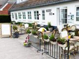 Udendørs opholdsområde i en hvid bygning med navnet Nordby Kro, med borde, stole, potteplanter, små græskar og danske flag på et dekorativt rækværk.