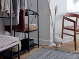 Et moderne, minimalistisk rum med en vævet stol, en sort metalhylde med foldede tekstiler og kurve, et tekstureret tæppe og en dekorativ vase med tørret græs på et trægulv.