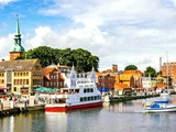 Farverig havnefront med både, der ligger til kaj, folk, der går langs promenaden, og charmerende europæiske bygninger, herunder en kirke med et grønt spir, under en delvist overskyet himmel.