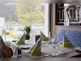 Et restaurantbord dækket til fire med foldede servietter, vinglas og en vase med gule blomster ved et vindue med udsigt til træer og en flod.