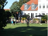 En mand putter på en green på en golfbane med en hvid bygning, udendørs siddepladser og mennesker i baggrunden.