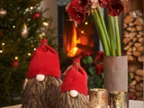 To nissefigurer med røde hatte sidder ved siden af guldlysestager og en vase med røde blomster med et tændt juletræ og en pejs i baggrunden.
