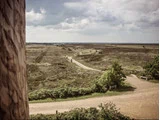 En snoet grussti løber gennem et græsklædt, åbent landskab under en overskyet himmel med spredte buske og to bænke nær stiens krydsningspunkt.