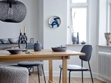 En moderne spisestue med træbord, sorte stole, keramisk service og minimalistisk indretning i neutrale toner. Lyset strømmer ind gennem de høje vinduer og lyser rummet op.