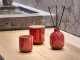 Tre røde keramikbeholdere står på en lys træoverflade; en krukke med låg, en åben kop og en vase med sorte sivpinde.