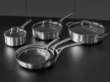 Et sæt køkkengrej i rustfrit stål, bl.a. tre stegepander og tre gryder med låg, anbragt på en sort bordplade.
