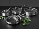 Et sæt gryder og pander i rustfrit stål med låg på en sort overflade, med et par stykker broccoli og rosenkål i nærheden.