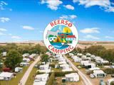 Luftfoto af en stor campingplads med campingvogne og træer, med et cirkulært Reersø Camping-logo over scenen.
