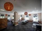 En rummelig, solbeskinnet restaurant med store vinduer, mørke stole, hvide duge og runde orange hængelamper.