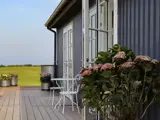En hyggelig gårdhave med et hvidt metalbord og -stole, hortensiaer i potter og et stribet gråt hus med udsigt over en grøn mark under en klar himmel.