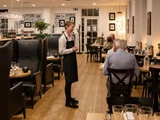 En tjener iført sort forklæde står ved et bord og taler til to siddende gæster i en varmt oplyst, moderne restaurant med flere optagede borde.