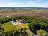 Luftfoto af en stor hvid bygning med rødt tag omgivet af træer, parkeringsplads og store åbne marker under en klar blå himmel.