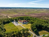 Luftfoto af en stor hvid bygning med rødt tag omgivet af træer i et stort, åbent landskab under en klar blå himmel.