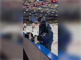 En ishockeymålmand i blåt udstyr står ved banden med vandflasker; i baggrunden træner spillere i hvidt og rødt på isen.