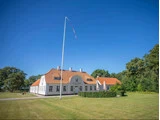 Et stort hvidt hus med rødt tegltag ligger på en velplejet græsplæne under en klar blå himmel med en høj, tom flagstang foran og træer i baggrunden.