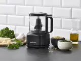 En sort Nordic Sense foodprocessor står på et køkkenbord omgivet af basilikum, hvidløg, parmesan, pinjekerner, pesto og olivenolie med hvide metrofliser i baggrunden.