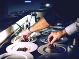 En kok anretter gourmetretter og placerer omhyggeligt maden på flere hvide tallerkener, der står på rad og række på en disk i et svagt oplyst køkken.