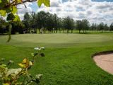 To personer står på en golfgreen omgivet af træer, med en sandbunker i forgrunden og en overskyet himmel over sig.