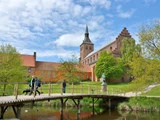 Folk går over en træbro over en dam med en historisk murstenskirke og træer i baggrunden på en delvis overskyet dag.