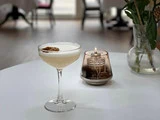 En skummende cocktail pyntet med en tørret citrusskive står på et hvidt bord ved siden af en lille olielampe i glas og en vase med grønne blade.