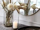 En glasvase med hvide tørrede blomster og græsser står på et træbord ved siden af et tændt stearinlys i en glasholder, og et rundt spejl reflekterer arrangementet.