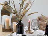 Et bord med tørret græs i en mørk vase, stearinlys i metallanterner, et tændt fyrfadslys i en holder og et vinglas på en linneddug.