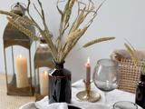 Et bord med tørret græs i en mørk vase, stearinlys i metallanterner, et tændt fyrfadslys i en holder og et vinglas på en linneddug.