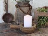 Et tændt hvidt bloklys står i en rustik træskål på et træbord, omgivet af lerkrukker og grønne planter.