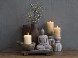 En rolig scene med en lille buddhastatue, tre tændte stearinlys, rustikke trævaser med bær og en morter og støder på en træoverflade mod en struktureret grå væg.