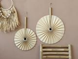 To runde, håndvævede palmevifter med bambushåndtag hænger på en beige væg ved siden af et dekorativt stykke strå og muslingeskaller; en bambusbænk står nedenunder.