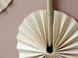 Nærbillede af en dekorativ håndvifte lavet af plisseret, lyst materiale med et bambushåndtag, sat op mod en dæmpet rosa tekstureret væg.