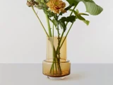 En gul-orange glasvase rummer et par grønne stilke og en gul blomst, der er placeret på en lys overflade mod en hvid baggrund.