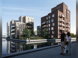 To personer går langs en sti ved vandet i nærheden af moderne boligblokke med altaner og store vinduer, træer og roligt vand, der reflekterer bygningerne.