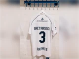 En hvid AGF Aarhus-fodboldtrøje med navnet Gretarsson, nummer 3 og et Andelskassen-logo, der hænger på et vægstativ.