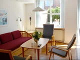 En lys stue med en rød sofa, to lænestole, et træbord med en blomstervase og sollys, der strømmer ind gennem et vindue, hvor man ser to mennesker udenfor.