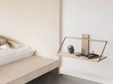 Et minimalistisk soveværelse med et svævende natbord af træ, hvorpå der står en lille højttaler, briller, en pen og en telefon, ved siden af en seng med hvidt sengetøj.