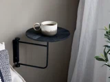 Et keramikkrus med kaffe står på et lille, rundt, væghængt sort bord ved siden af en beige væg, tæt på et gennemsigtigt gardin og en grøn plante.