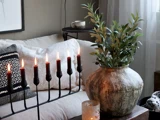 En hyggelig stue med en tændt sort menorah, mønstrede puder i en beige sofa, en stor rustik vase med grønne planter og et stearinlys i en glasholder på et træbord.