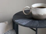 Et keramikkrus fyldt med sort kaffe står på et mørkt, struktureret sidebord ved siden af en pude med et sort og hvidt vævet mønster.