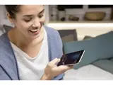 En kvinde smiler, mens hun holder en smartphone og taler ind i den, mens hun sidder indendørs i en sofa med puder i baggrunden.
