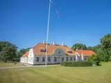 Stort hvidt hus med rødt tegltag og kvistvinduer på en grøn græsplæne med høj flagstang og træer under en klar blå himmel.