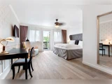 Lyst hotelværelse med dobbeltseng, skrivebord, stol, tv, lænestol, stort spejl og altandør, der lukker naturligt lys ind.