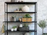 Sort metalhylde mod en betonvæg, dekoreret med potteplanter, keramikvaser, kurve og en kasse med grønt for et rustikt, minimalistisk look.