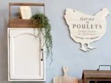 Et hvidt skab med en plante og en bog på toppen er monteret på en blå væg ved siden af et dekorativt fransk skilt i form af en kylling og skærebrætter i træ.