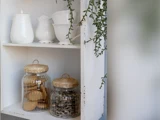 Hvid hylde med hvide keramikskåle og kander på toppen og to glaskrukker med flettede låg, den ene fyldt med småkager og den anden med frø, med en grøn plante hængende over.