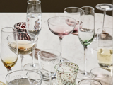 En række tomme og halvfyldte glas, herunder vin-, champagne- og vandglas, er anbragt på et lyst bord. Nogle glas har farvede eller strukturerede mønstre.