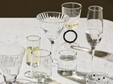 Forskellige klare glas med forskellige former og struktureret design er anbragt på en lys overflade, og hvert glas indeholder vand eller hvidvin.