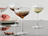 Fire elegante glas på stilk med indviklede mønstre, hver især fyldt med forskellige drikkevarer, er anbragt på en reflekterende overflade i nærheden af nogle spredte mandler og stablede tallerkener.